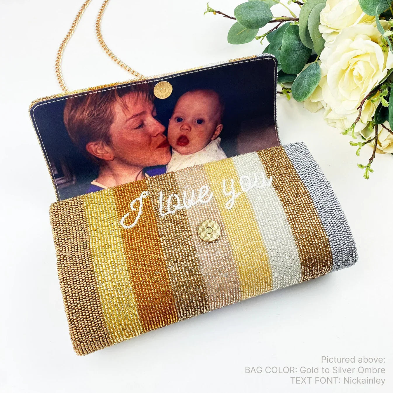 A handbag with a photo of a woman and a baby inside, along with the text "I love you" printed on it. The bag has a striped pattern with gold and silver beads. There is also a bouquet of white flowers in the background.