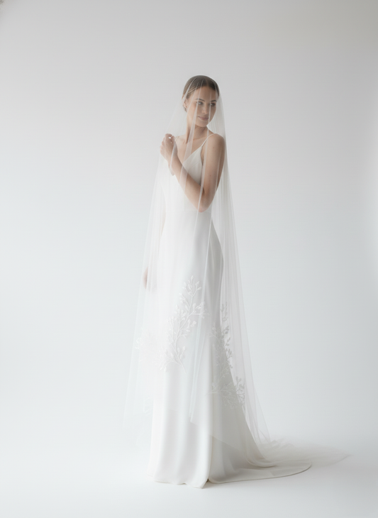 A woman in a long white dress stands in a studio, her face partially obscured by a veil.