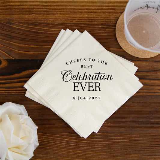 Stack of napkins with 'Cheers to the best celebration ever' text on a wooden table with flowers and a glass.