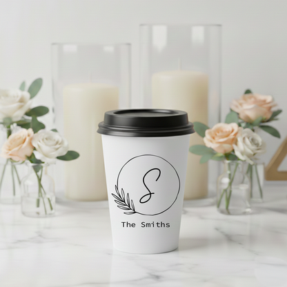 A white coffee cup with a black lid and a logo featuring a letter "S" is placed on a marble surface, surrounded by vases of flowers and candles.
