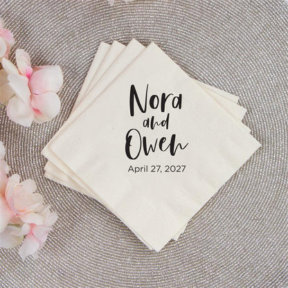 Stack of personalized napkins with 'Nora and Owen' and date on a textured surface with flowers.