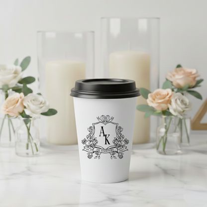 A white coffee cup with a black lid, featuring a monogrammed logo, is placed on a marble surface. In the background, there are two tall glass vases with white candles inside them, and several small vases containing pink roses.
