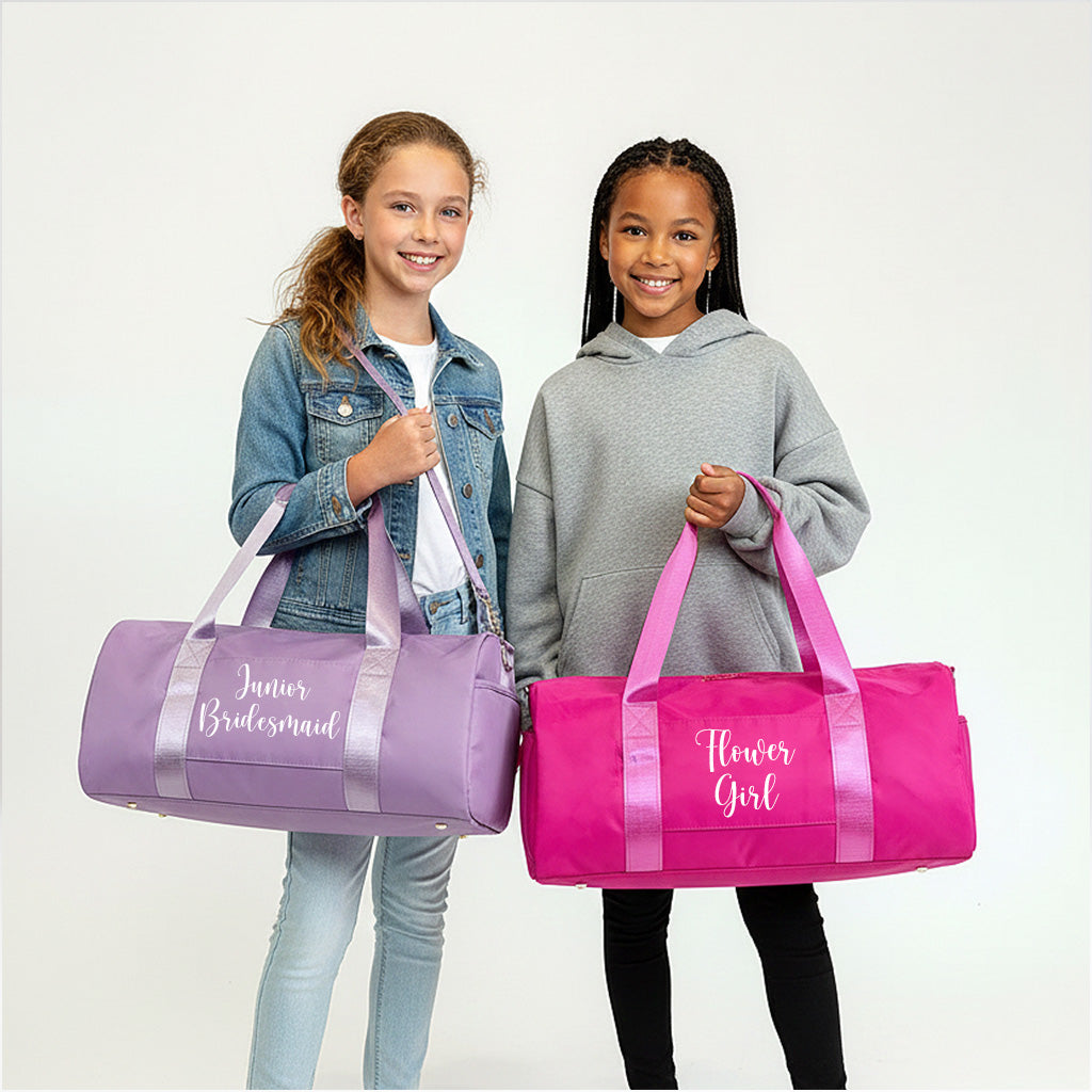 Two young girls are standing next to each other, each holding a bag. The girl on the left is holding a purple bag with the text "Junior Bridesmaid" written on it, while the girl on the right is holding a pink bag with the text "Flower Girl" written on it.