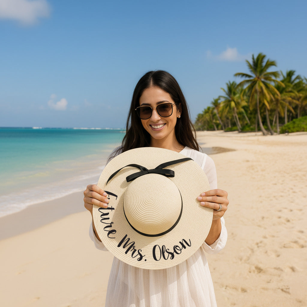 A woman stands on a beach holding a large white hat with the text "Future Mrs. Olson" written on it.