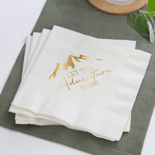 Stack of white napkins with gold text 'Let the Adventure Begin' on a green surface.