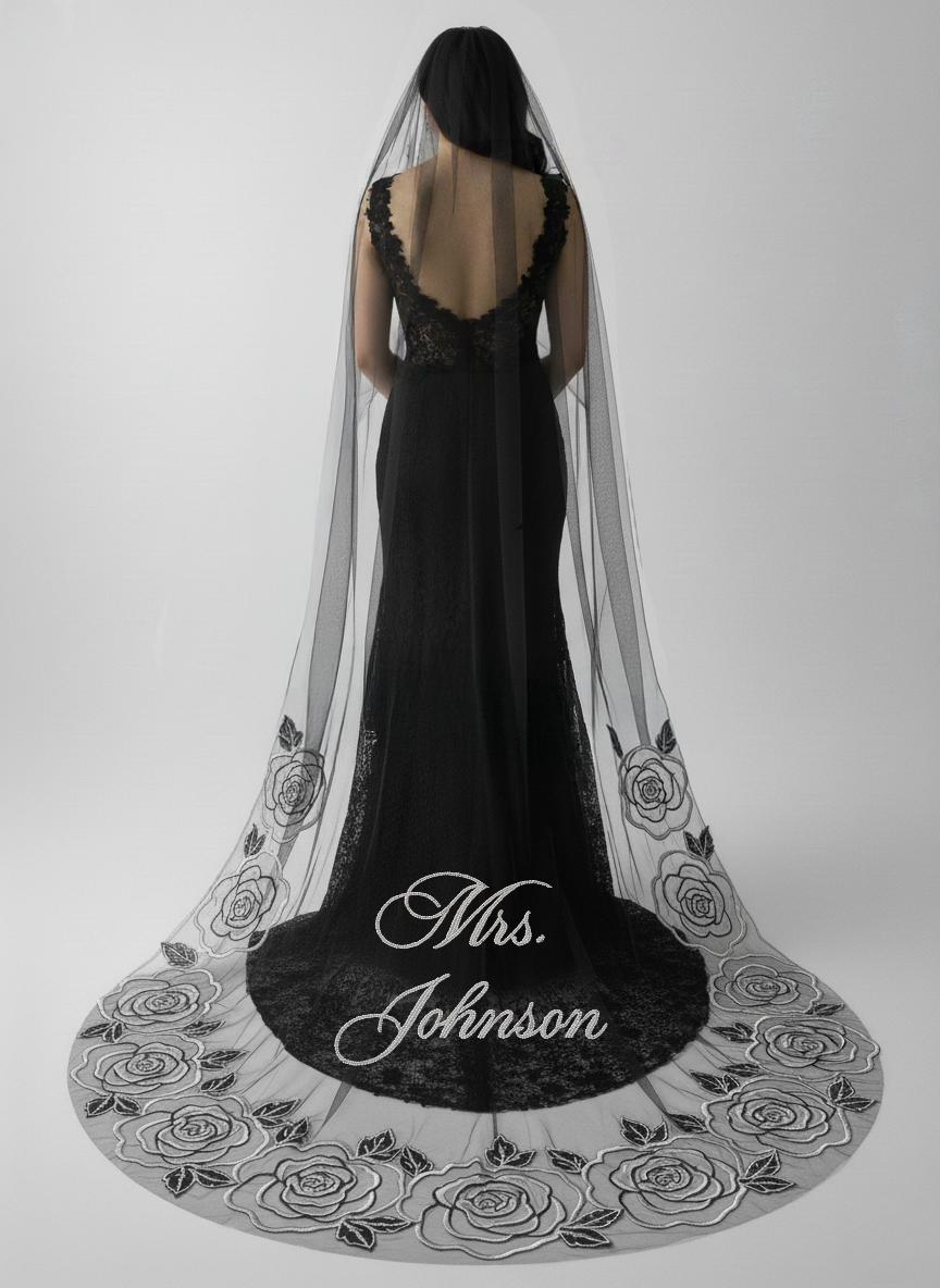 A woman wearing a black lace dress with a long, flowing veil decorated with black roses stands against a plain white background.