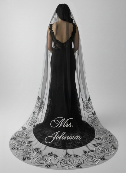 A woman wearing a black lace dress with a long, flowing veil decorated with black roses stands against a plain white background.