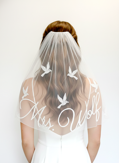 A woman wearing a white wedding dress with a veil featuring the name 'Mrs. Wolf' and a design of white birds.