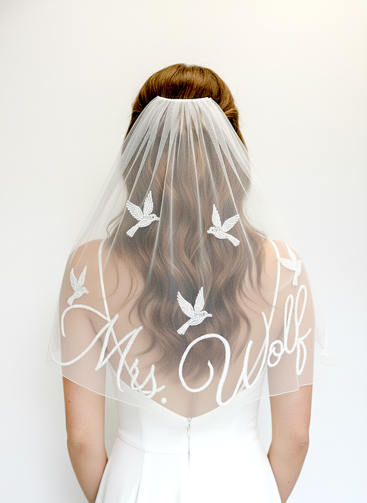 A woman wearing a white wedding dress with a veil featuring the name 'Mrs. Wolf' and a design of white birds.