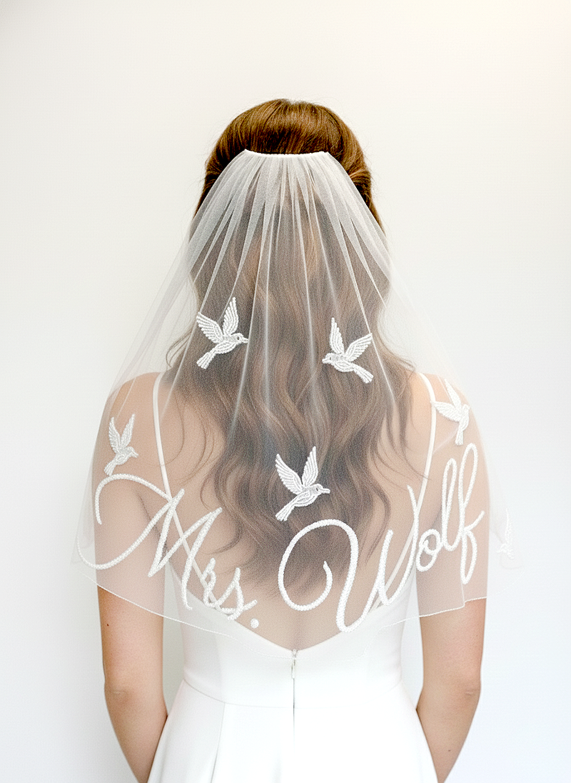 A woman wearing a white wedding dress with a veil featuring the name 'Mrs. Wolf' and a design of white birds.