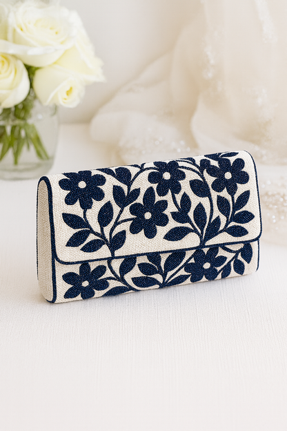 A navy blue and white clutch purse with a floral pattern is displayed on a white surface, with a vase of white roses in the background.