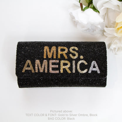 A black clutch purse with the text "MRS. AMERICA" embroidered on it, placed on a white surface next to a bouquet of white flowers.