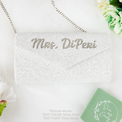 A white clutch purse with the text "Mrs. DiPiperi" embroidered on it, placed on a white surface next to a green box and some flowers.