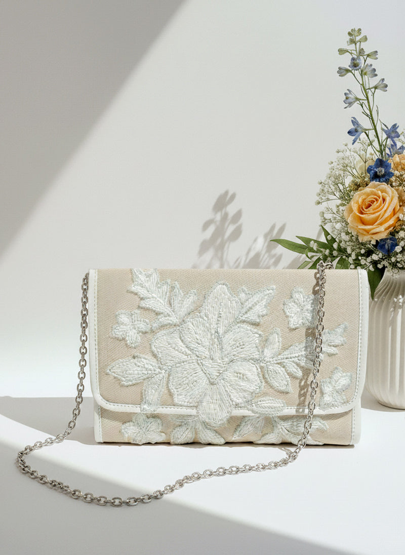 A beige clutch purse with a floral embroidered design sits on a white surface, accompanied by a vase of flowers.