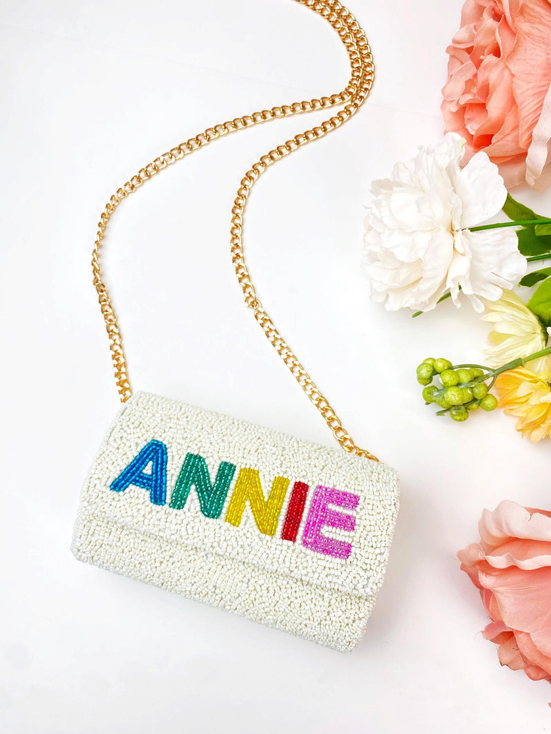 A white handbag with the name "ANNIE" embroidered on it, placed on a white background next to some pink flowers.