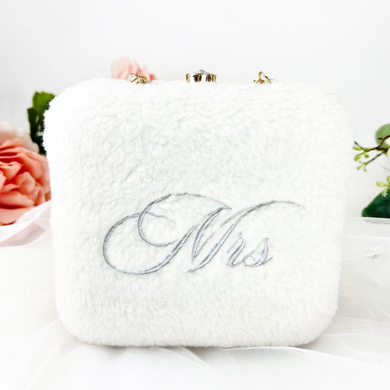 A white fur clutch bag with the word "Mrs" embroidered on it, placed on a white surface with pink flowers in the background.