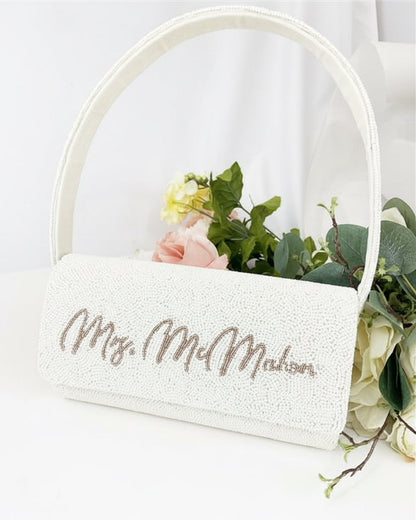 A white handbag with the text "Mrs. M. M. M." embroidered on it, placed on a white surface next to a bouquet of flowers.