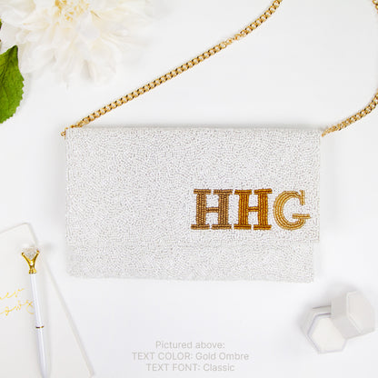 a white clutch purse with the initials "HHG" embroidered on it, placed on a white surface alongside a pen, a flower, and some hexagonal shapes.