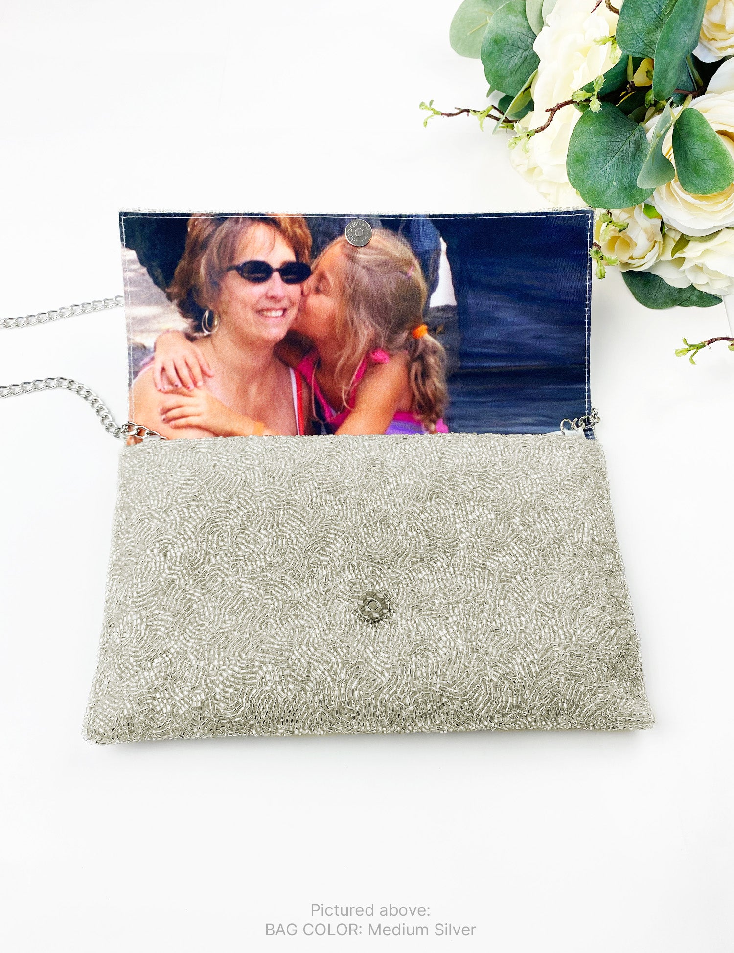 a silver clutch purse with a photo of two women inside, one of whom is kissing the other. The purse is placed on a white surface, and there are some flowers visible in the background.