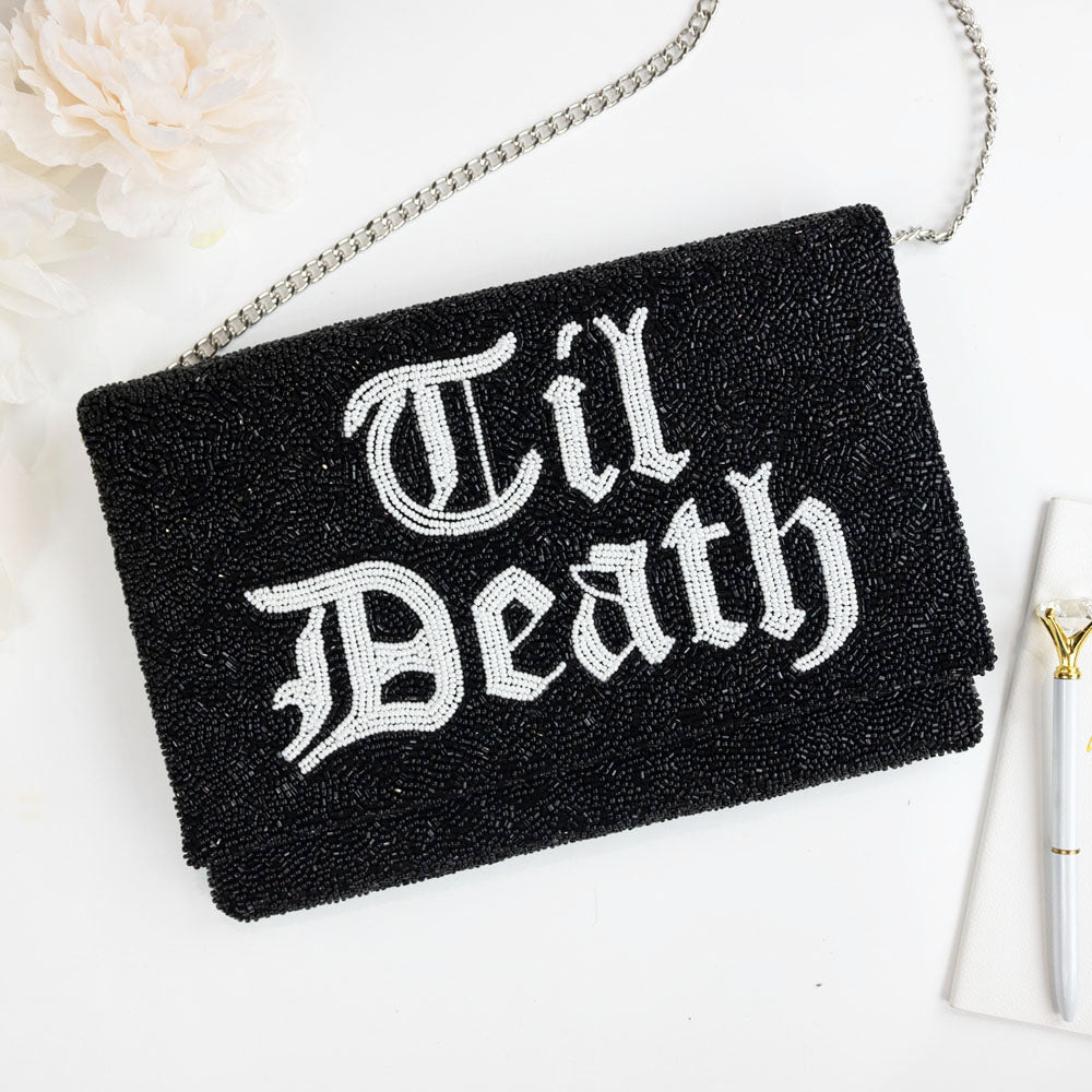 A black clutch purse with the text "Til Death" embroidered on it, placed on a white surface next to a white flower.