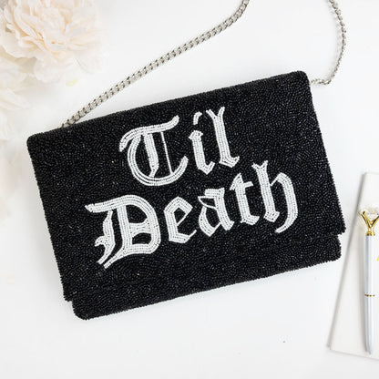 A black clutch purse with the text "Til Death" embroidered on it, placed on a white surface next to a white flower.