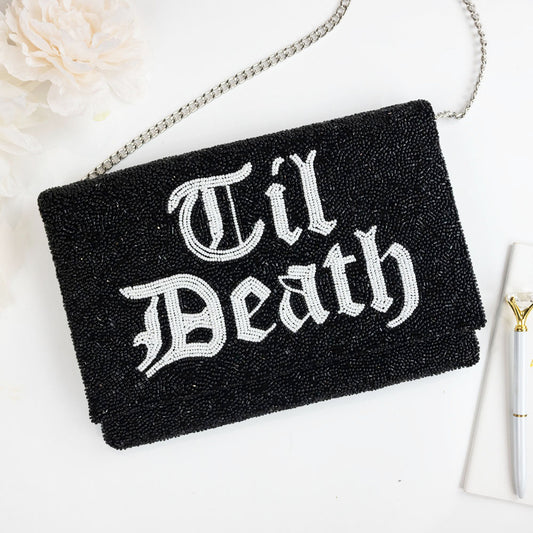 A black clutch purse with the text "Til Death" embroidered on it, placed on a white surface next to a white flower.