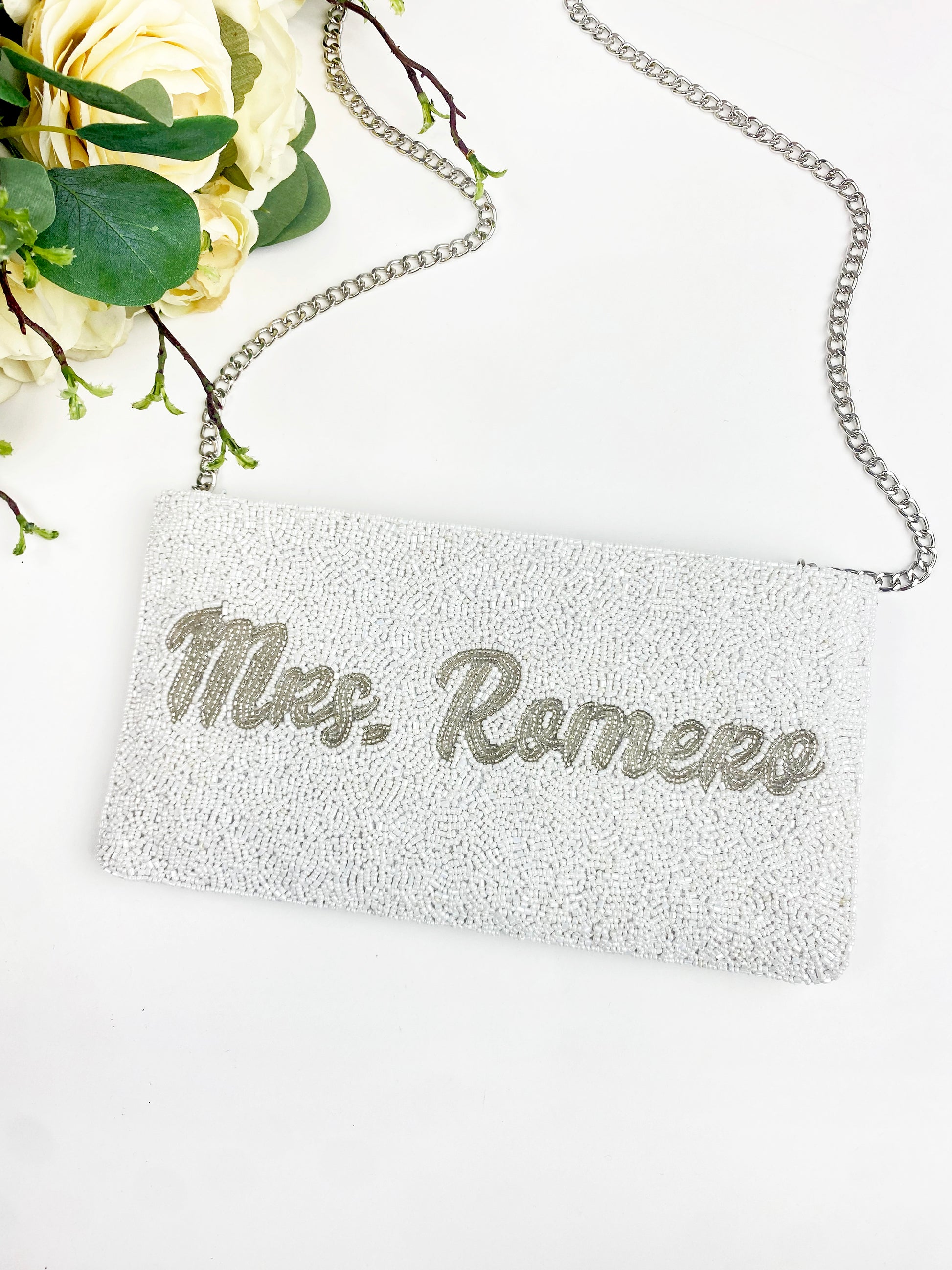 A white clutch purse with the name "Mrs. Romero" printed on it, placed on a white surface next to a bouquet of yellow flowers.