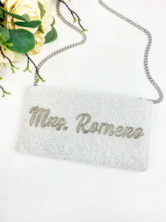 A white clutch purse with the name "Mrs. Romero" printed on it, placed on a white surface next to a bouquet of yellow flowers.
