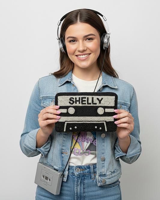 A young woman wearing headphones and a denim jacket is holding a black and white cassette tape bag with the word "SHELLY" printed on it.