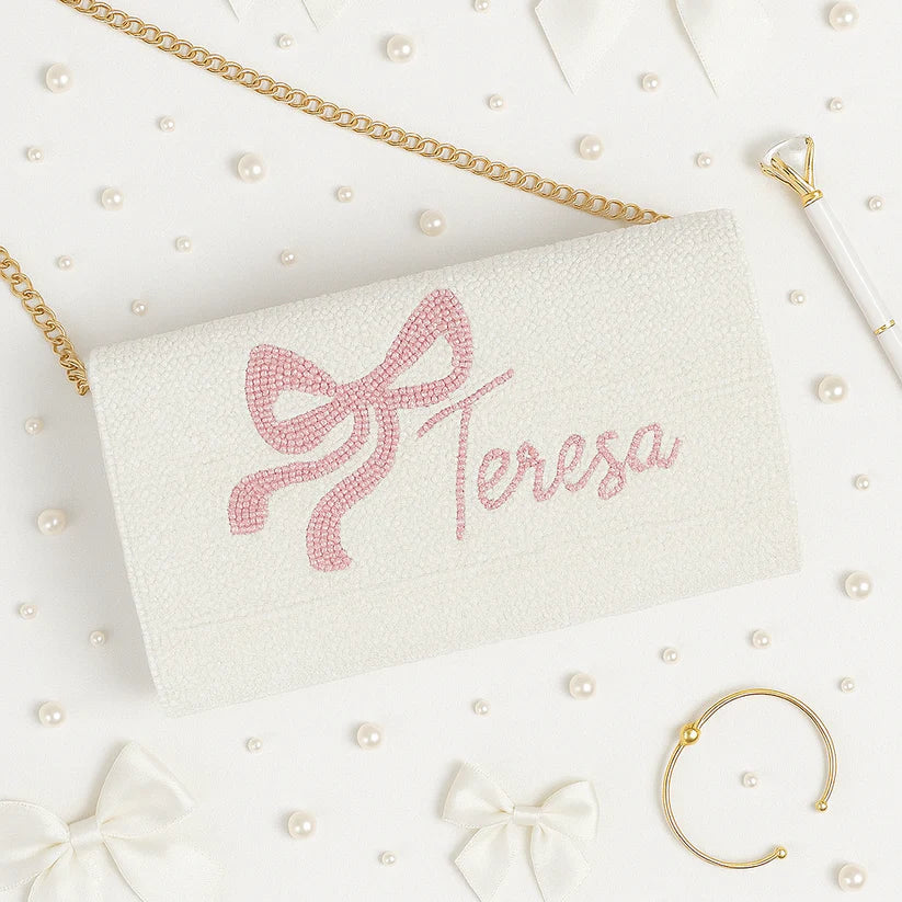 a white clutch purse with a pink bow and the name "Tresa" embroidered on it, surrounded by various accessories such as pearls, a bracelet, and a pen.