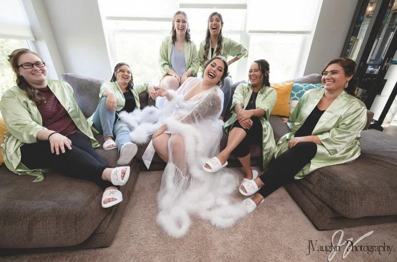 Bride in white feather robe laughing with bridesmaids wearing matching sage green satin robes — fun bridal party getting-ready photo