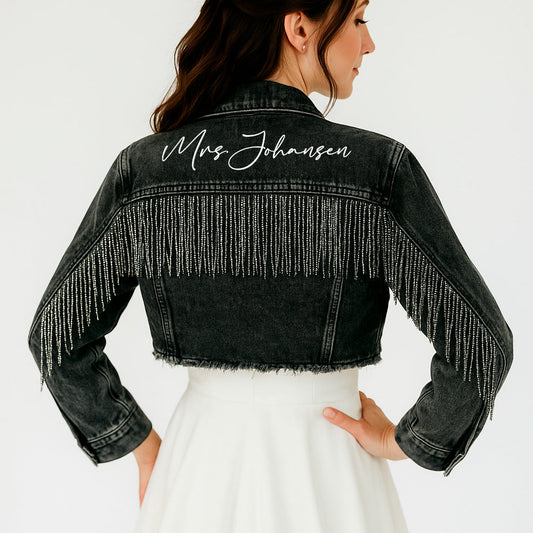 Black denim jacket with rhinestone fringe and 'Mrs. Johansen' embroidery on a white background