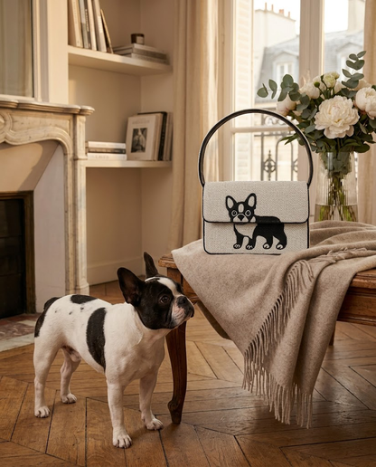 A small black and white dog stands on a wooden floor, looking at a handbag with a dog design on it, which is placed on a wooden table.