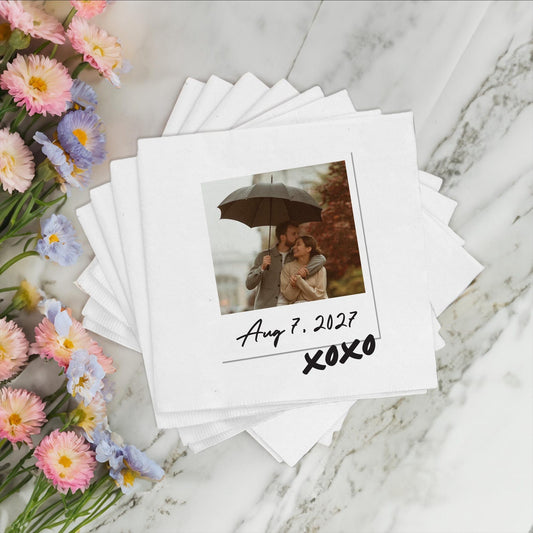 Stack of napkins with photo prints of a couple under an umbrella, dated August 7, 2027, on a marble surface with flowers.