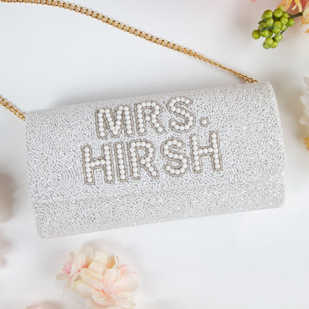 A white clutch purse with the text "MRS. HIRSH" printed on it, surrounded by flowers and a gold chain strap.