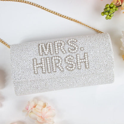 A white clutch purse with the text "MRS. HIRSH" printed on it, surrounded by flowers and a gold chain strap.