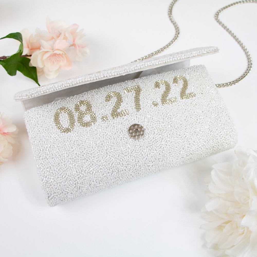 "Handmade pearl beaded bridal clutch with an intricate pattern, perfect for wedding day essentials. Comes with a gold or silver chain, sized at 9.5in x 5in x 3in. Features the option for a custom date inside. Unique design variations make each bridal clutch one-of-a-kind."