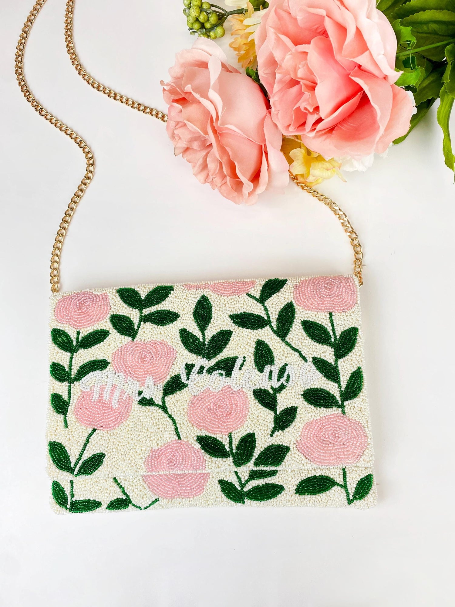 a white clutch purse with a floral pattern and a chain strap, placed next to a bouquet of pink flowers.